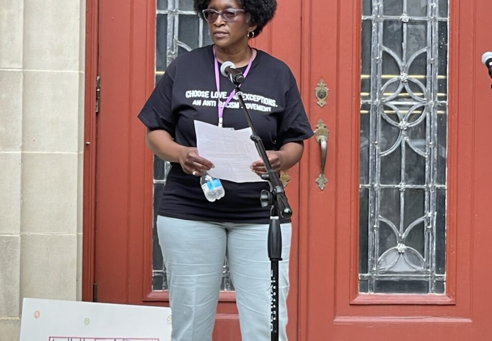 A person stands outside a building, holding a sign with the text "CHOOSE LOVE. EXCEPTIONS. AN ANTI-RACISM MOVEMENT. UNLEARN RACISM. 613" and a human face on it.
