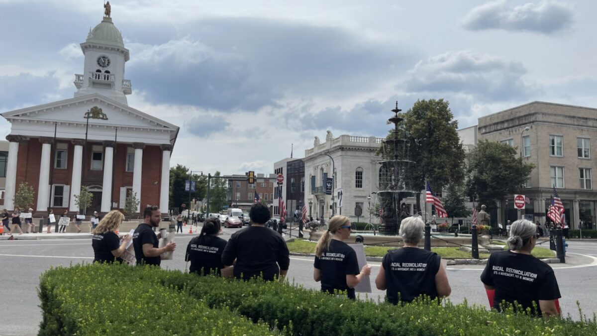 People of all ages and backgrounds are gathering in the city's government building, surrounded by trees, grass, plants, and a church, while a cloudy sky looms overhead and a busy street runs nearby.