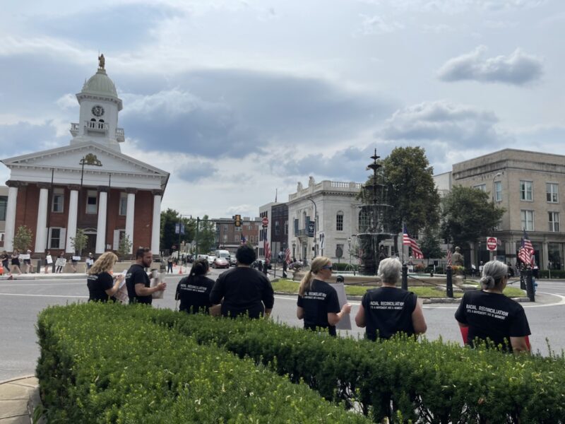 People of all ages and backgrounds are gathering in the city streets around a government building, surrounded by trees, grass, plants, and a cloudy sky.