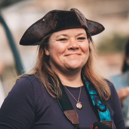 Courtney Robertson, an open source developer relations advocate, is wearing a pirate hat, a DSLR camera, and a purple shirt