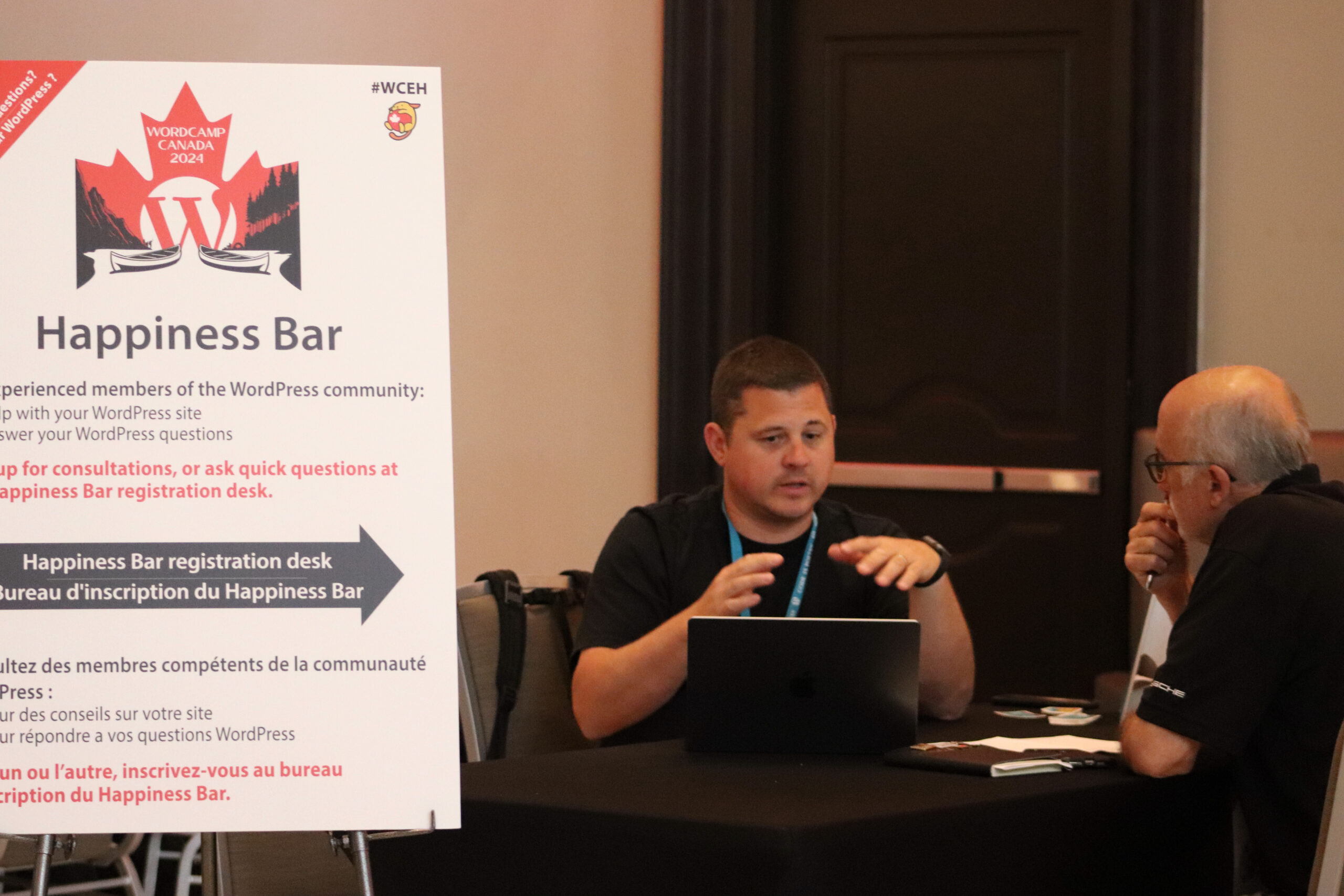 Ryan Welcher sits at WordCamp Canada's Happiness Bar with a laptop in front of him at the table. Across from him is a bald man with his hands to his lips in deep thought. 