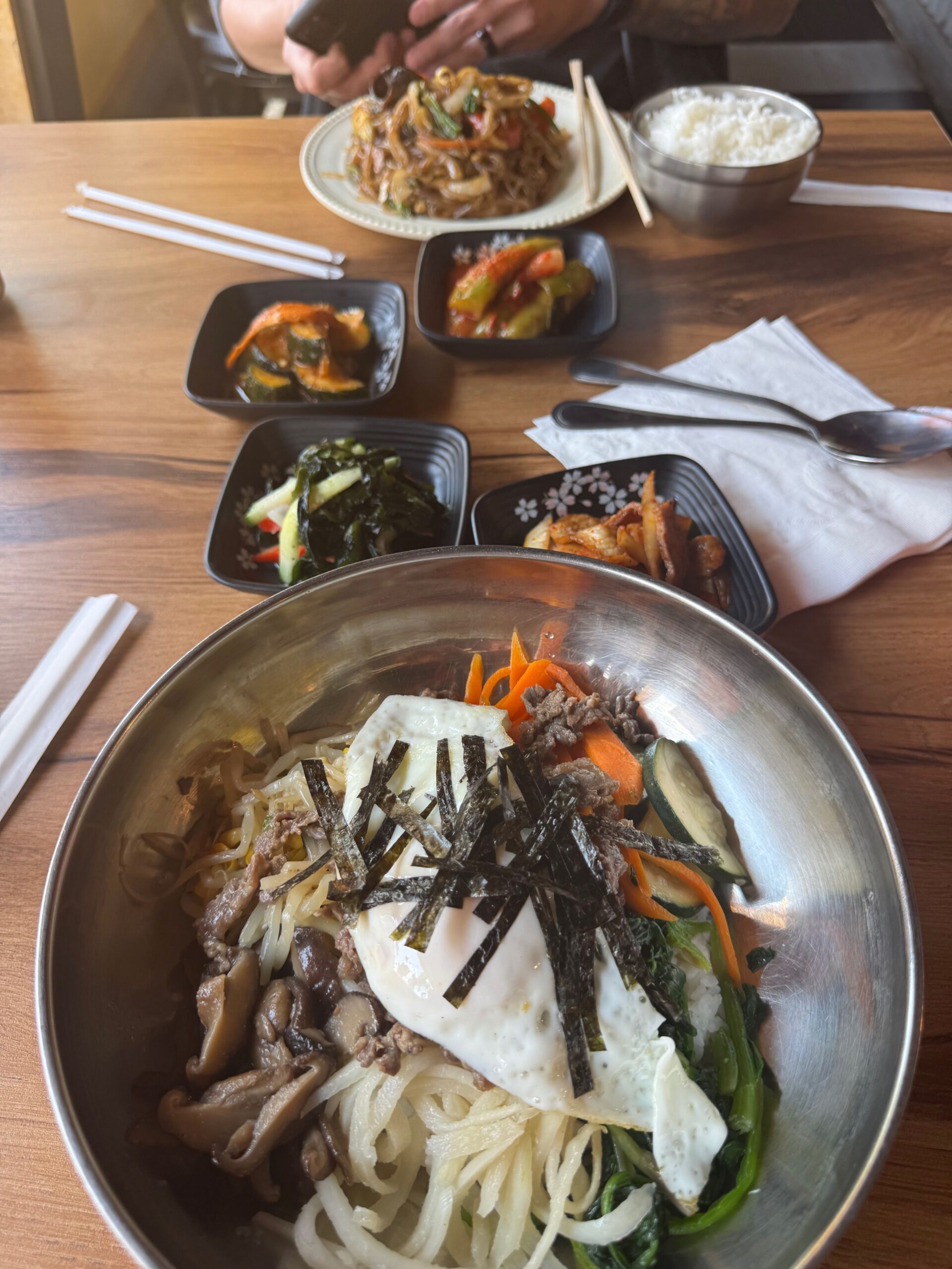 
A meal setup on a wooden table with two main dishes and several side dishes. In the foreground, there is a bowl of bibimbap with vegetables, mushrooms, and a fried egg on top, garnished with shredded seaweed. In the background, a person is holding a phone above a plate of stir-fried noodles with vegetables. Surrounding the main dishes are small bowls containing various banchan (Korean side dishes) including kimchi, pickled vegetables, and a bowl of rice. Chopsticks and a spoon are placed next to the dishes.