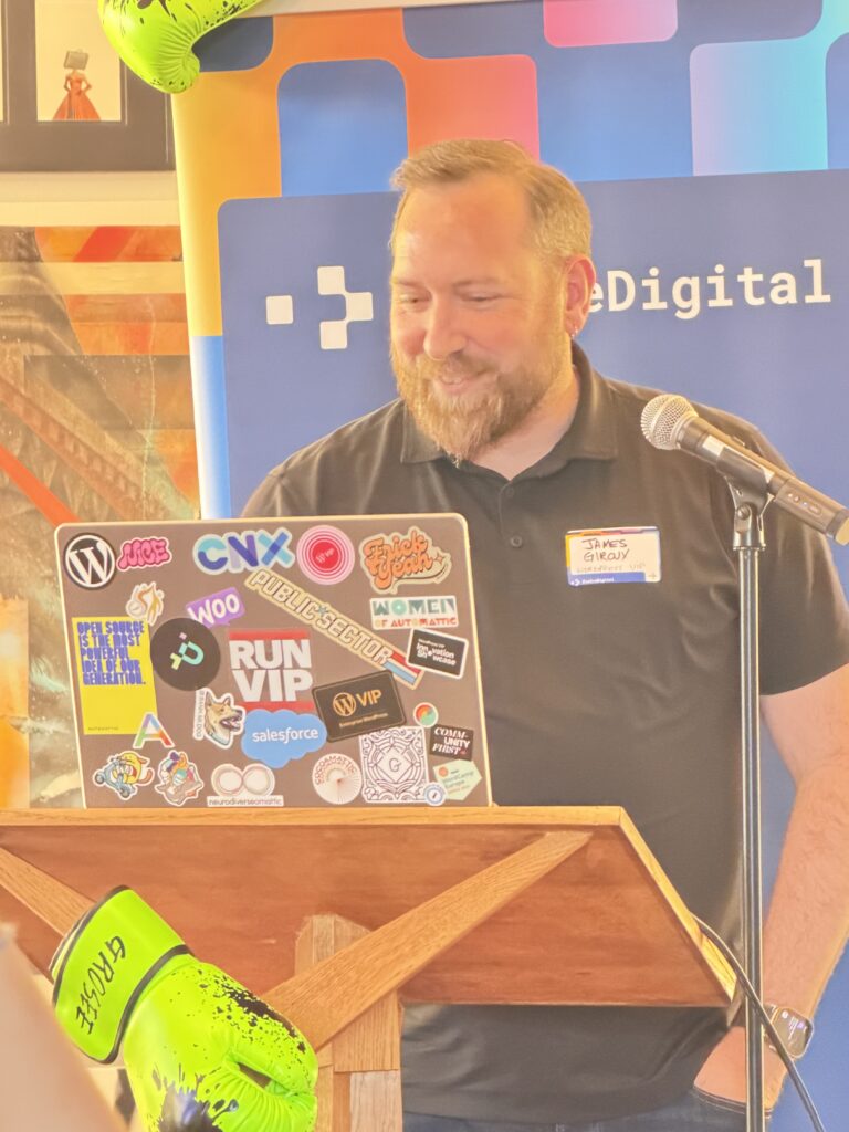 James Giroux of WordPress VIP smiles at a sticker-covered laptop on a podium framed by neon green boxing gloves