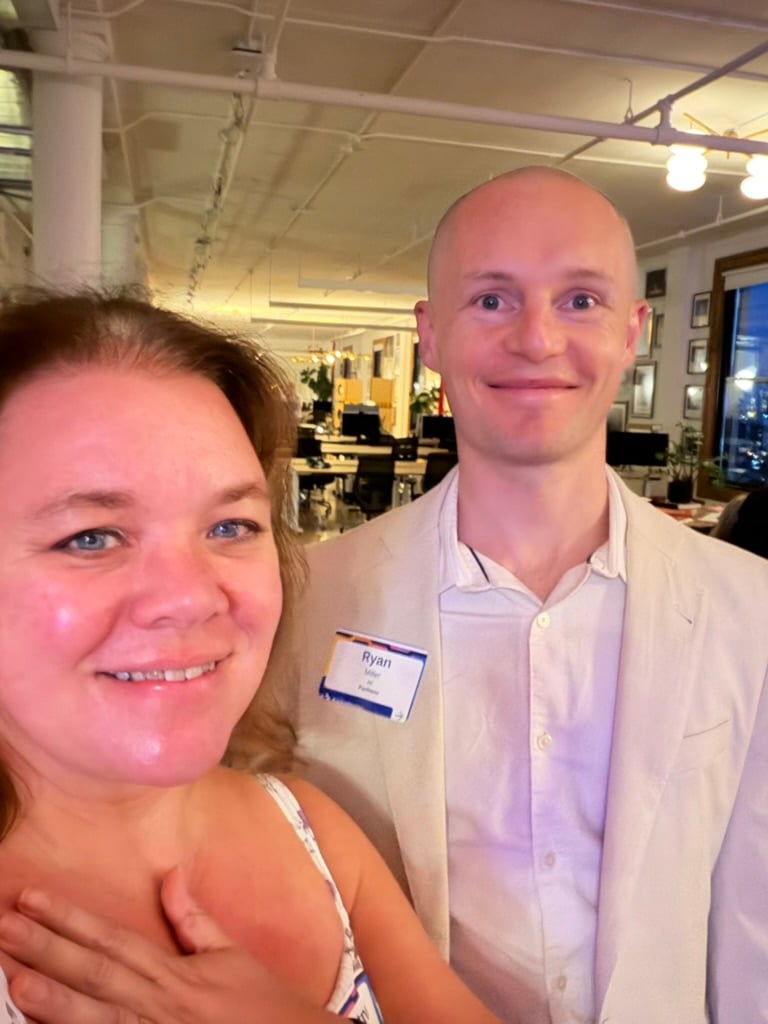Courtney smiles for a selfie beside Ryan, who wears a light blazer and name tag, inside a warmly lit office event space.