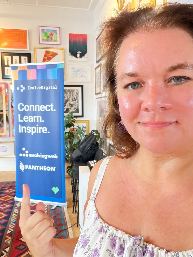 Courtney in a white floral dress smiles and points toward an EvolveDigital banner reading “Connect. Learn. Inspire.” with Evolving Web and Pantheon logos, surrounded by colorful framed artwork. She is especially pointing to the Pantheon logo as a sponsor at the bottom of the banner.