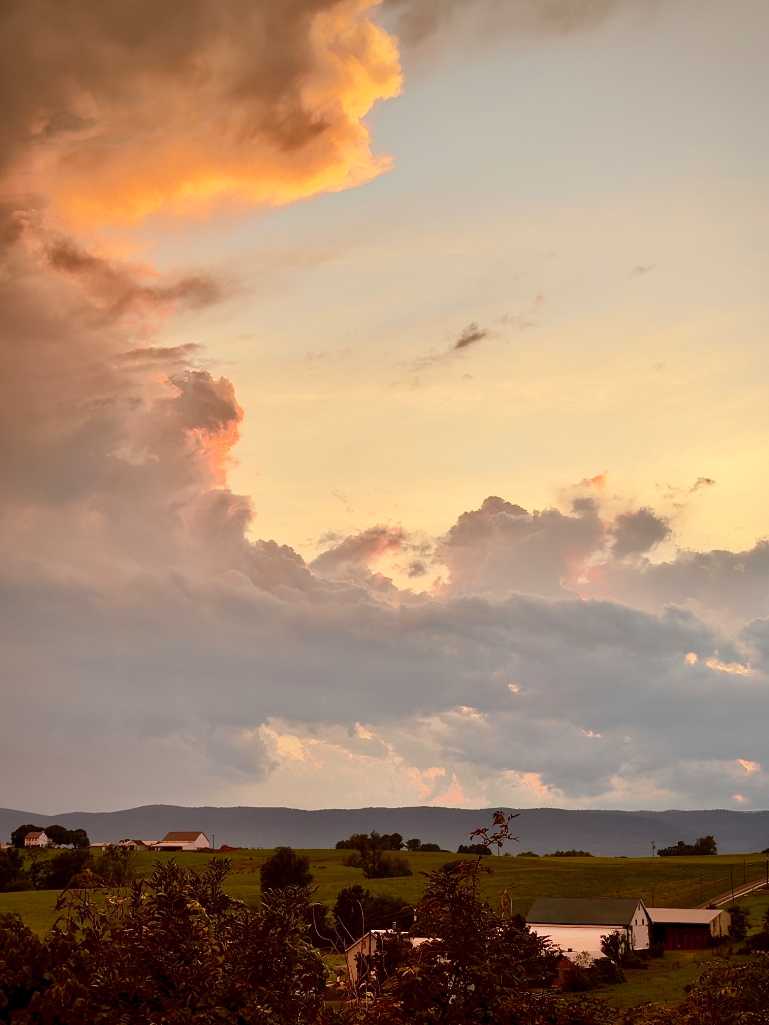 Orange sunset light illuminates towering storm clouds above green farmland, white barns, and a distant mountain ridge.