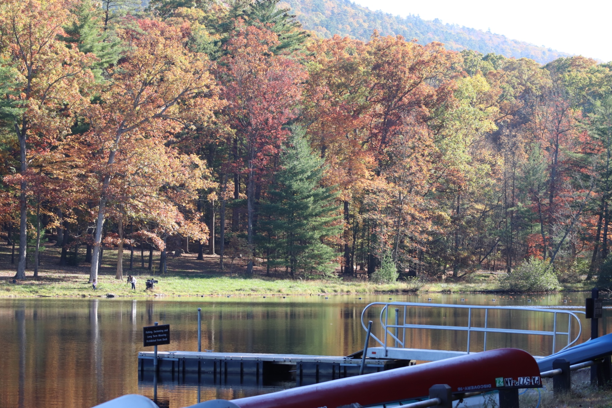 Small floating dock extending into a still lake, with a red canoe in the foreground and a dense forest of golden, orange, and red autumn trees across the water.